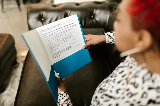 A woman sits on a couch reading a document in a blue folder. Business insurance selection checklist: verify the FCA Register, compare documents and record decisions