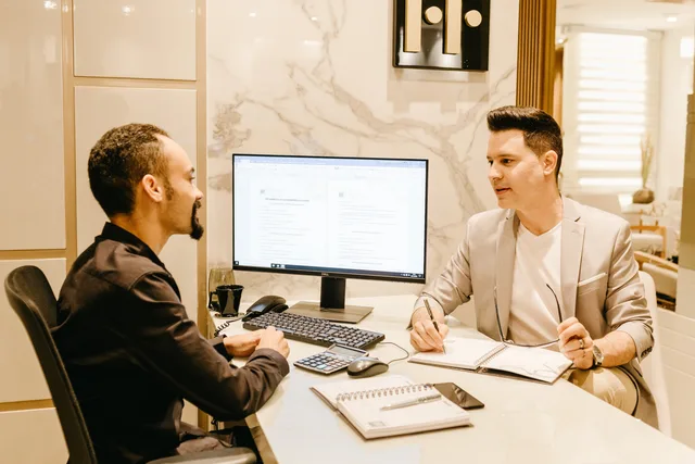 Two businessmen discussing work at a sleek and modern office desk, using computer and notes for collaboration. Business insurance software selection for SME brokers