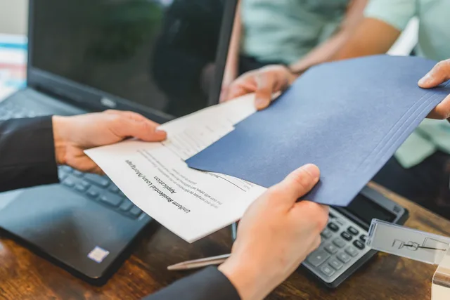 Close-up of hands exchanging real estate documents in an office setting. Eight business insurance roles to name, from executive risk owner to claim lead